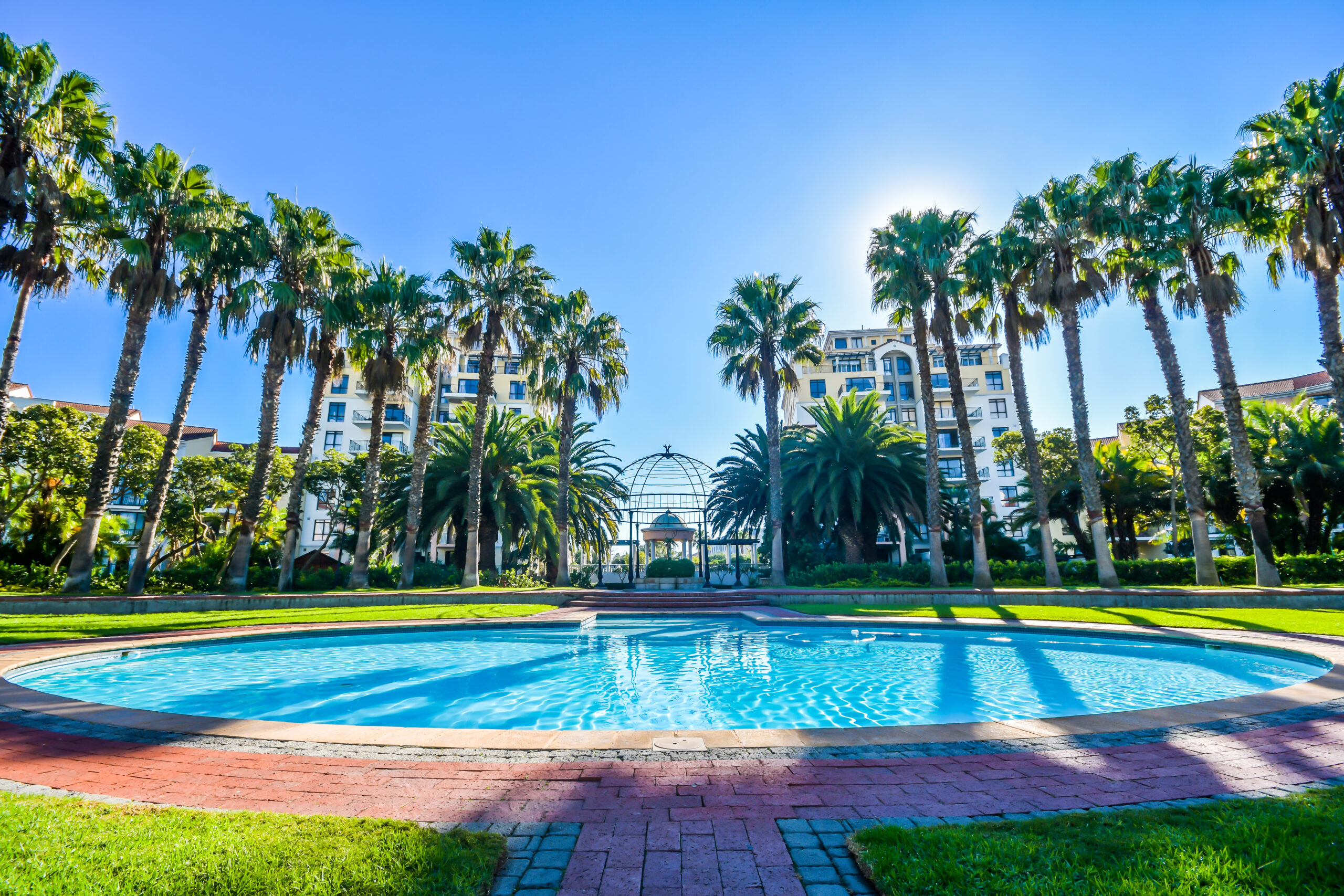 A round swimming pool surrounded by palm trees, with tall apartment buildings in the background under a bright blue sky—perfect for a Cape Town city stay with 20% OFF.