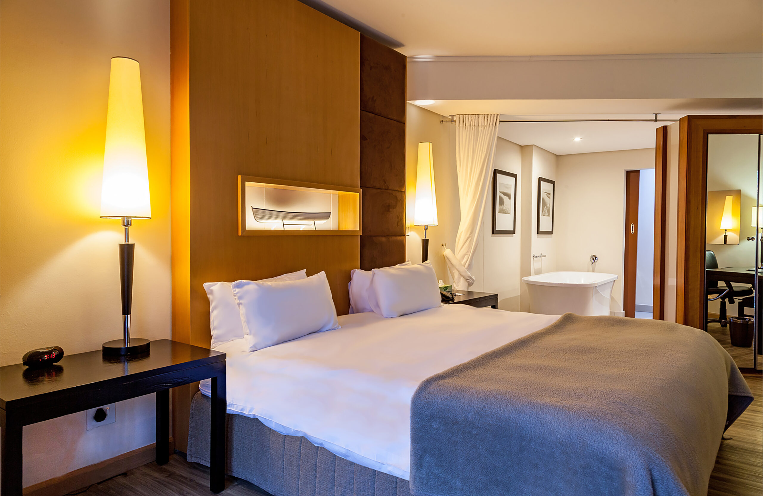 A modern hotel room with a large bed, white linens, two tall lamps, a bedside table, a gray blanket, framed art on the wall, and a freestanding bathtub in the background—perfect for guests seeking comfort or those looking to take advantage of our exclusive pensioner special.