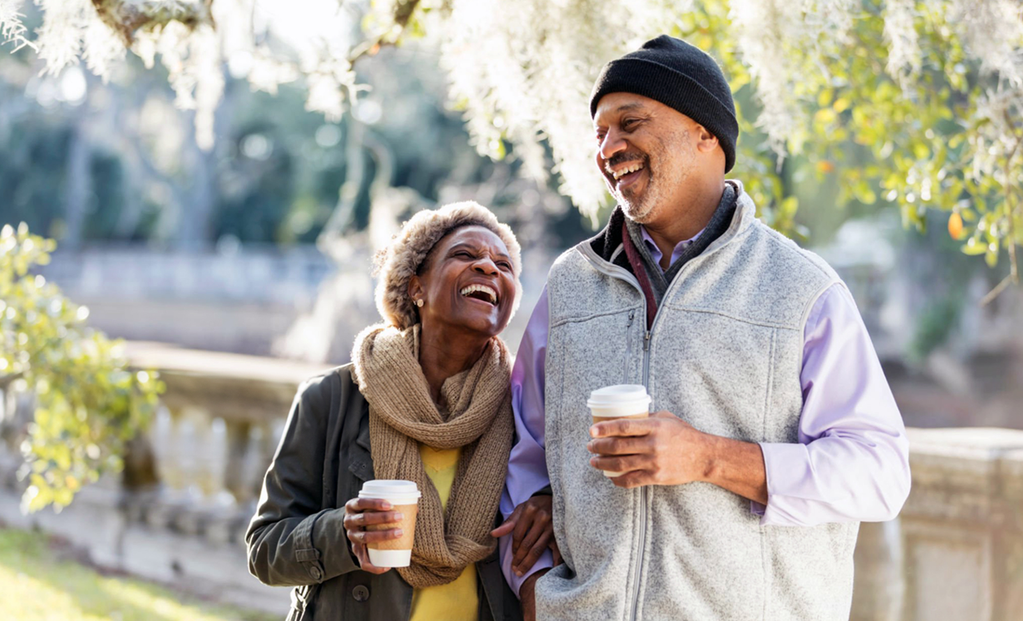 An older couple smiles and laughs while walking outdoors, each holding a coffee cup. The woman wears a scarf and jacket, and the man wears a beanie and vest. Sunlight filters through trees in the background, capturing the joy of spending quality time together—just like you can during your Cape Town city stay with 20% OFF.